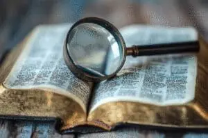 An open, aged Bible with a magnifying glass on its table of contents, focusing on chapter counts per book